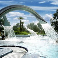 Ein Au&szlig;enpool mit Wasserfont&auml;nen und Palmen, umgeben von B&auml;umen und einem Geb&auml;ude. Menschen genie&szlig;en das Wasser unter blauem Himmel., &copy; Stuttgarter B&auml;der