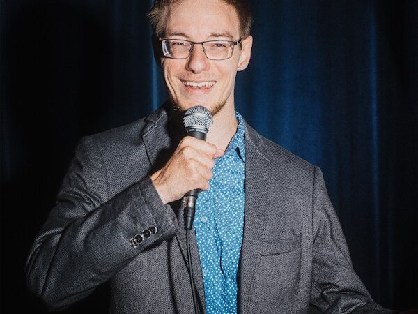 Ein Mann mit Brille und Mikrofon steht l&auml;chelnd vor einem blauen Vorhang. Er tr&auml;gt ein graues Sakko und ein blaues Hemd., &copy; Rosenau Kultur e.V.