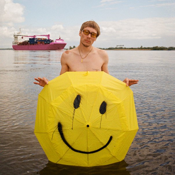 Ein Mann mit Sonnenbrille steht im Wasser und hält einen gelben Schirm mit Smiley. Im Hintergrund fährt ein großes Schiff vorbei., © Rosenau Kultur e.V. Ein Mann mit Sonnenbrille steht im Wasser und hält einen gelben Schirm mit Smiley. Im Hintergrund fährt ein großes Schiff vorbei., © Rosenau Kultur e.V.
