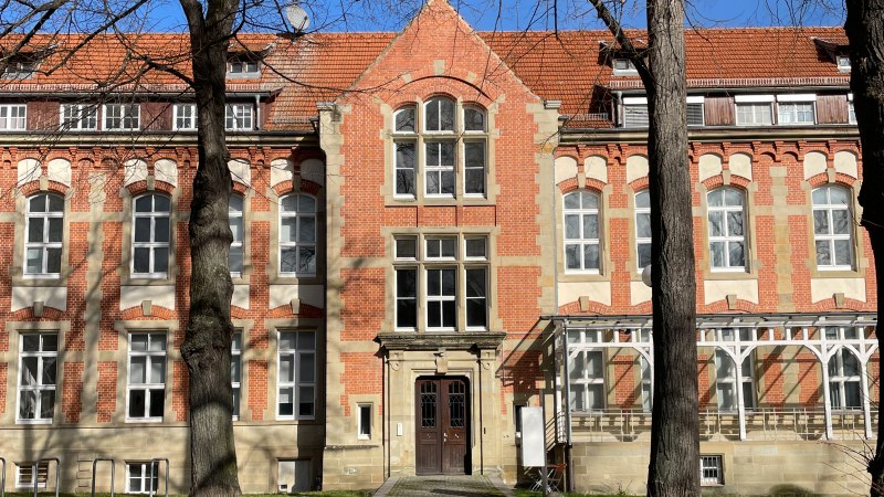 Ein historisches Geb&auml;ude aus rotem Backstein mit gro&szlig;en Fenstern und einem Giebeldach, umgeben von B&auml;umen und einem blauen Himmel., &copy; Merz Berufskolleg f&uuml;r Grafik-Design