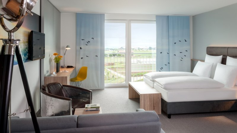 Modernes Hotelzimmer mit großem Bett, Schreibtisch, Sessel und Balkon mit Aussicht auf Landschaft. Dekor mit blauen Vorhängen und Vogelmotiven., © Essential by Dorint Modernes Hotelzimmer mit großem Bett, Schreibtisch, Sessel und Balkon mit Aussicht auf Landschaft. Dekor mit blauen Vorhängen und Vogelmotiven., © Essential by Dorint