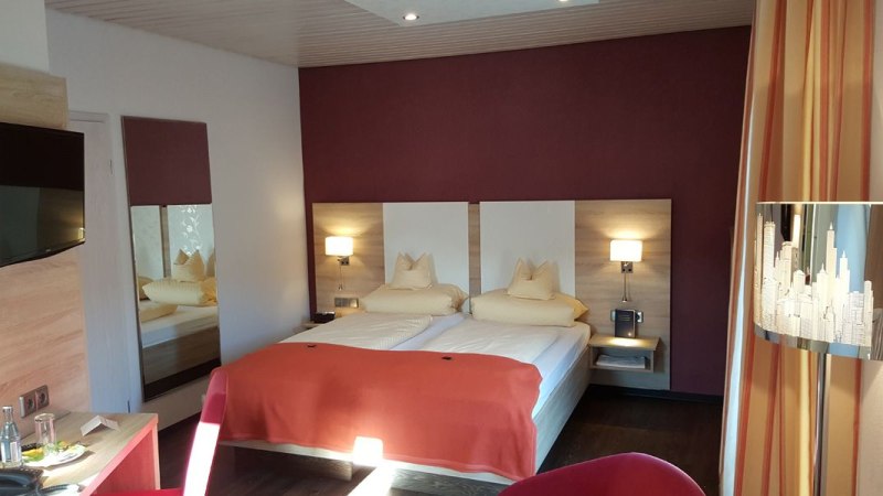 Modernes Doppelzimmer mit zwei Betten, roter Wand, Fernseher und stilvoller Beleuchtung. Ein Tisch mit Wasserflasche und Gläsern steht bereit., © Hotel Koch Modernes Doppelzimmer mit zwei Betten, roter Wand, Fernseher und stilvoller Beleuchtung. Ein Tisch mit Wasserflasche und Gläsern steht bereit., © Hotel Koch