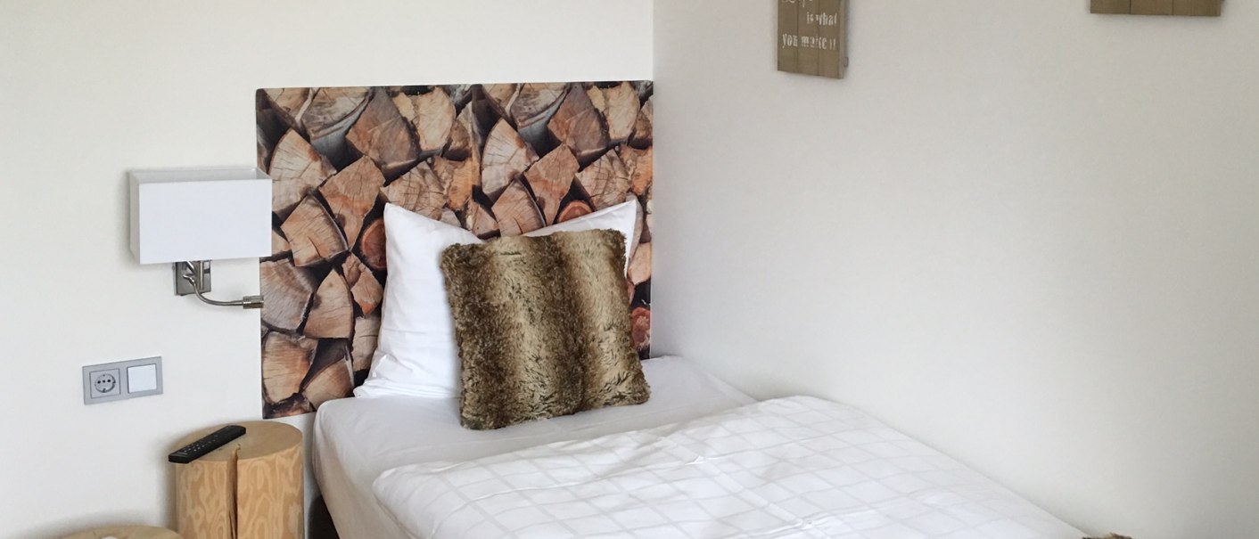 Ein gemütliches Einzelbett mit Holzoptik-Kopfteil, Fellkissen und Decke. An der Wand hängen motivierende Sprüche., © TOMAS
