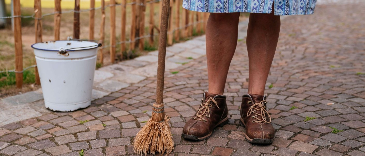 Person in braunen Schuhen steht auf Kopfsteinpflaster, hält einen Besen. Ein weißer Eimer steht daneben. Die Person trägt einen blauen Rock., © Thomas Niedermüller Person in braunen Schuhen steht auf Kopfsteinpflaster, hält einen Besen. Ein weißer Eimer steht daneben. Die Person trägt einen blauen Rock., © Thomas Niedermüller