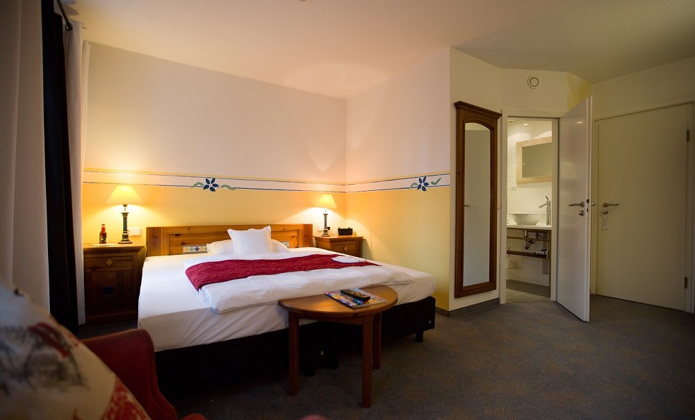 A cozy hotel room with a large bed, red accents, nightstands, lamps, and an open bathroom. A mirror hangs on the wall., &copy; Hotel B&uuml;rkle