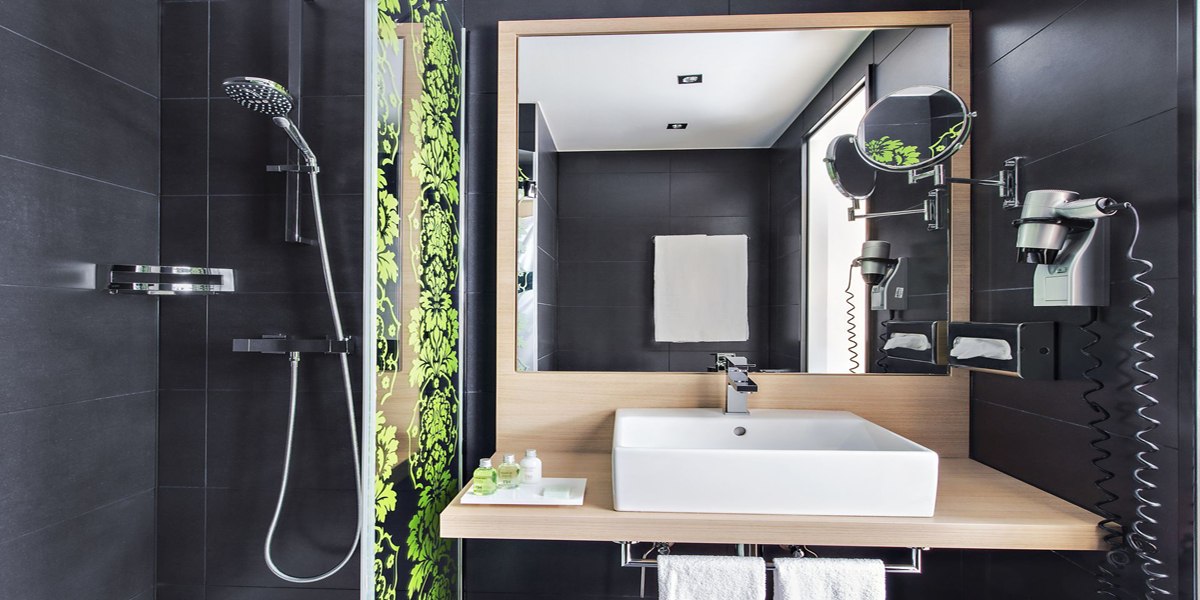 Modern bathroom with a black tiled wall, sink, large mirror, shower, and hair dryer. Green plant patterns serve as decoration., © NH Ludwigsburg Modern bathroom with a black tiled wall, sink, large mirror, shower, and hair dryer. Green plant patterns serve as decoration., © NH Ludwigsburg