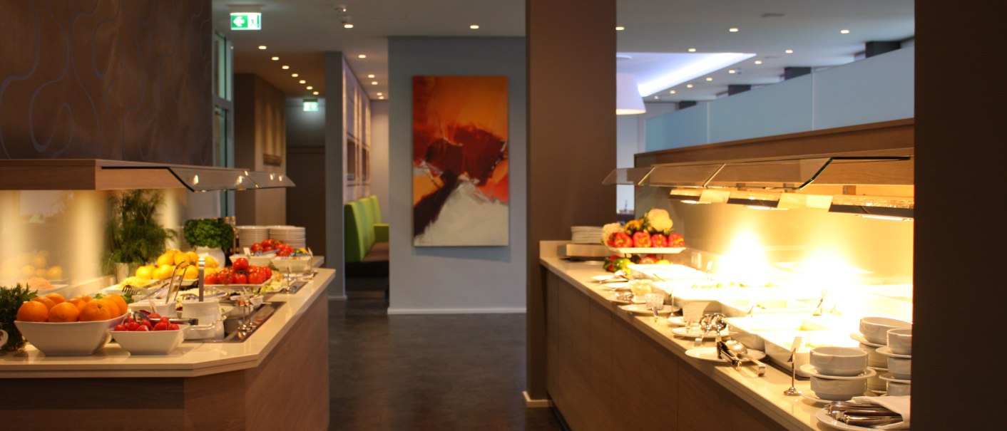 Ein stilvolles Buffet in einem Restaurant mit Obst, Geschirr und warmem Licht. Moderne Einrichtung mit Kunst an der Wand., © Hotel am Schlossberg Herrenberg