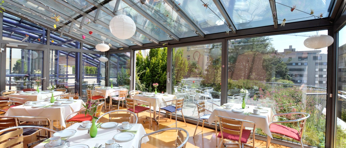 Ein lichtdurchfluteter Wintergarten mit gedeckten Tischen, Stühlen und Glaswänden, umgeben von Pflanzen. Ideal für ein Frühstück im Freien., © Abalon Hotel Ideal Ein lichtdurchfluteter Wintergarten mit gedeckten Tischen, Stühlen und Glaswänden, umgeben von Pflanzen. Ideal für ein Frühstück im Freien., © Abalon Hotel Ideal