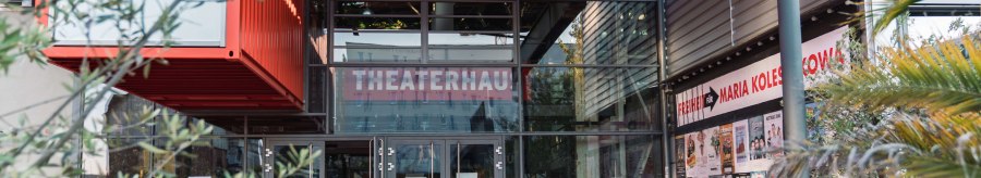 Entrance to the Theaterhaus Stuttgart with red floor, glass facade and posters. Two people enter the building. Plants and modern architecture are visible., © Stuttgart-Marketing GmbH, wpsteinheisser