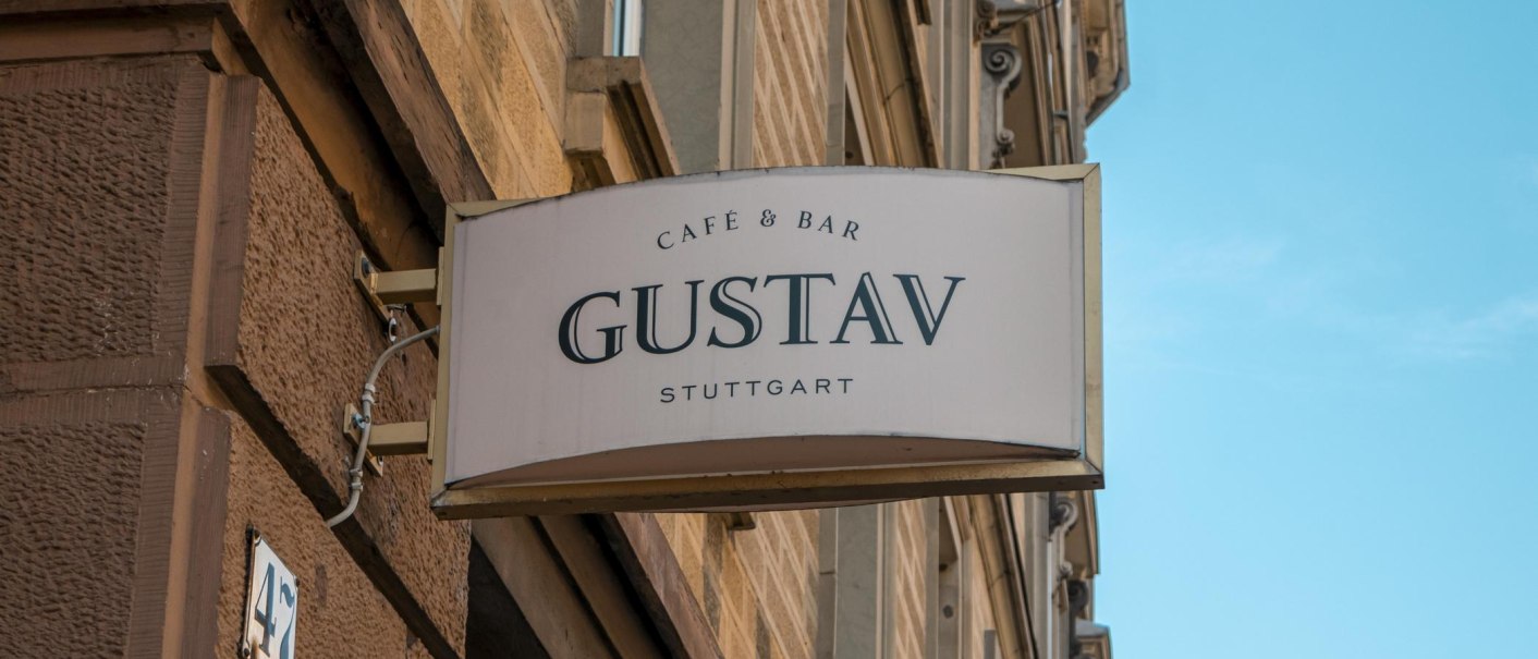 Ein Schild mit der Aufschrift 'Café & Bar Gustav Stuttgart' hängt an einer Hauswand. Der Himmel ist blau., © SMG, Sarah Schmid
