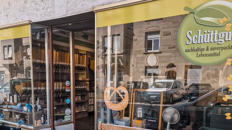 Außenansicht eines Unverpackt-Ladens mit Schüttgut-Logo. Im Schaufenster sind Behälter mit Lebensmitteln und ein Schild mit "BIO" zu sehen., © Schüttgut, Stuttgart