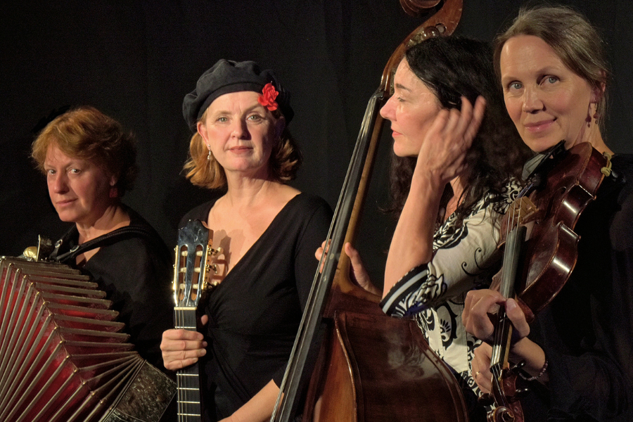 Vier Musikerinnen mit Akkordeon, Gitarre, Kontrabass und Geige posieren vor einem dunklen Hintergrund. Eine Frau tr&auml;gt eine schwarze Baskenm&uuml;tze mit roter Blume., &copy; Fotocredit Theater La Lune Stuttgart