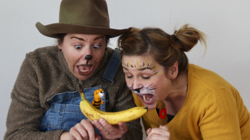 Zwei verkleidete Personen mit Tiergesichtsbemalung bestaunen eine Banane, auf der ein kleines Tiger-Spielzeug sitzt. Sie wirken überrascht und erfreut., © Theater Kopfkonfetti