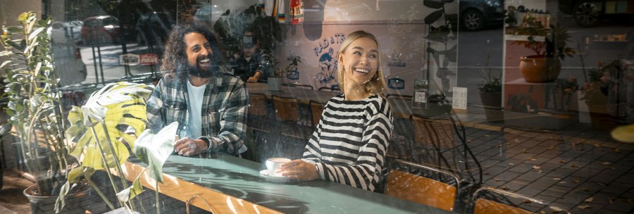 Zwei Personen sitzen l&auml;chelnd in einem Caf&eacute;, umgeben von Pflanzen. Sie genie&szlig;en eine entspannte Atmosph&auml;re, w&auml;hrend sie durch eine Glasscheibe betrachtet werden., &copy; SMG, Sarah Schmid