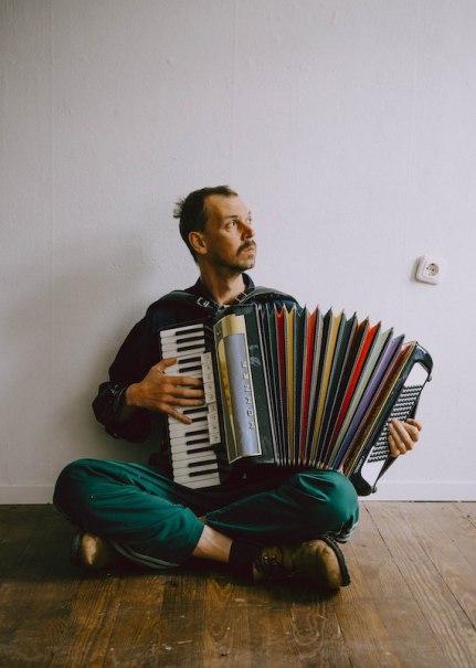 A man sits on the wooden floor and plays an accordion. He wears green pants and looks thoughtfully to the side., &copy; Rosenau Kultur e.V.