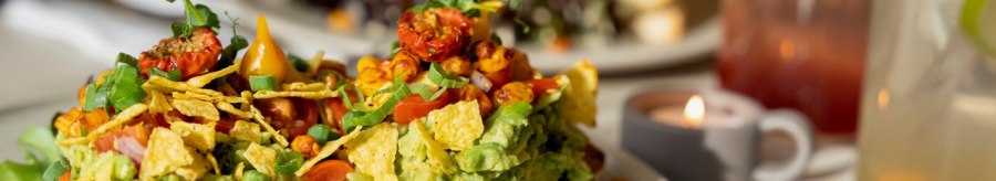 Avocado toast with tomatoes and herbs, a sandwich and drinks in the background. A tea light creates a cozy atmosphere., &copy; Veganes Gold&reg;