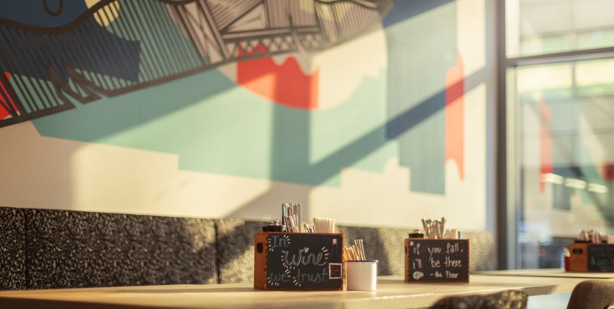 Ein Restaurant mit moderner Wandkunst und Tischen, auf denen Besteckbehälter stehen. Sonnenlicht fällt durch die Fenster., © Novum Hospitality