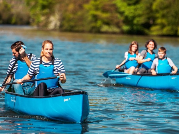 Two blue canoes with people paddling on a river. The people are wearing striped shirts and blue life jackets. There is green vegetation in the background., &copy; Cool-Tours StattReisen
