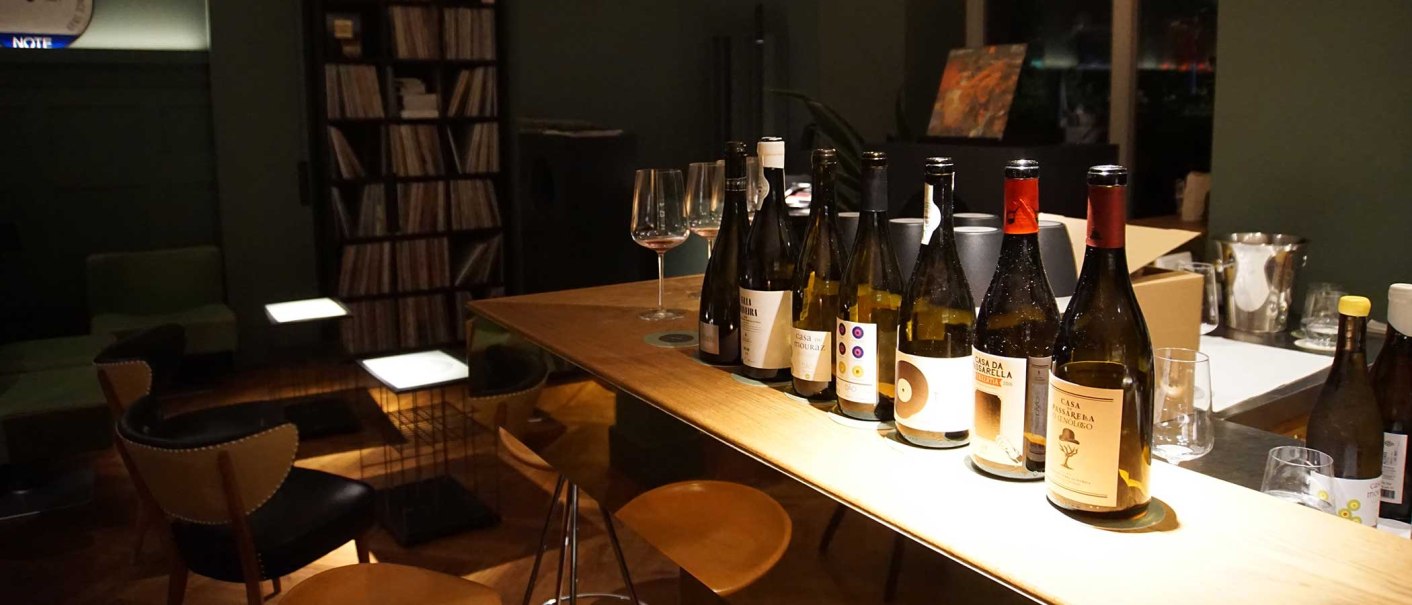 Several bottles of wine stand on a wooden table in a stylish bar. A record shelf and comfortable seating can be seen in the background., © Weinhandlung Kreis KG