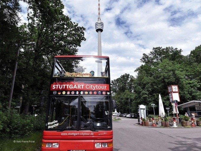 Red double-decker bus of the Stuttgart Citytour in front of the Stuttgart television tower, surrounded by trees and a café., © Willms Touristik Red double-decker bus of the Stuttgart Citytour in front of the Stuttgart television tower, surrounded by trees and a café., © Willms Touristik