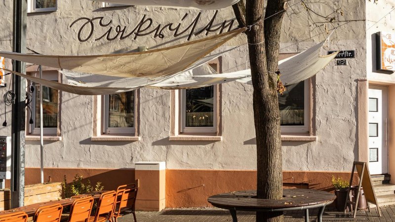 Ein Caf&eacute; mit Sonnensegeln und orangenen St&uuml;hlen im Au&szlig;enbereich. Die Fassade ist beige mit Schriftzug. Ein Baum steht vor dem Eingang., &copy; Stuttgart-Marketing GmbH, Sarah Schmid