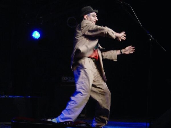 A man in a suit and hat gesticulates animatedly on a stage with a microphone, surrounded by dark lighting., &copy; Theaterhaus Stuttgart e.V.