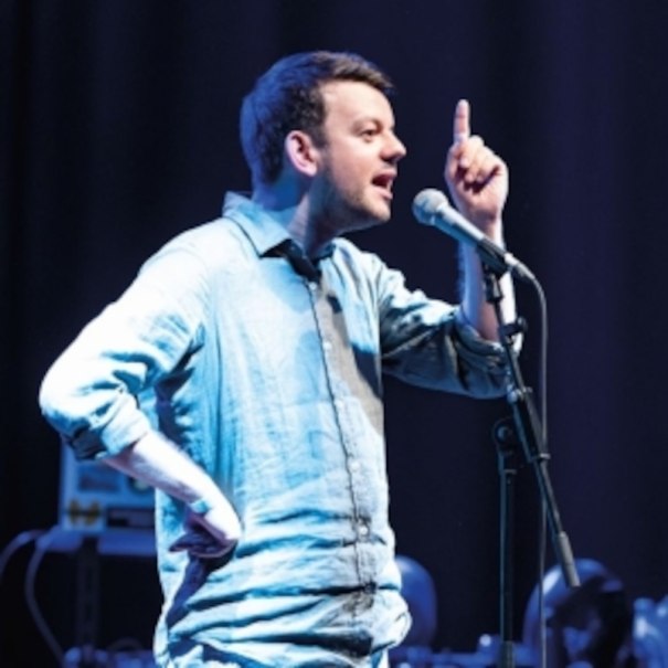 A man in a blue shirt gesticulates into a microphone on a stage., &copy; Altes Schauspielhaus und Kom&ouml;die im Marquardt e.V.