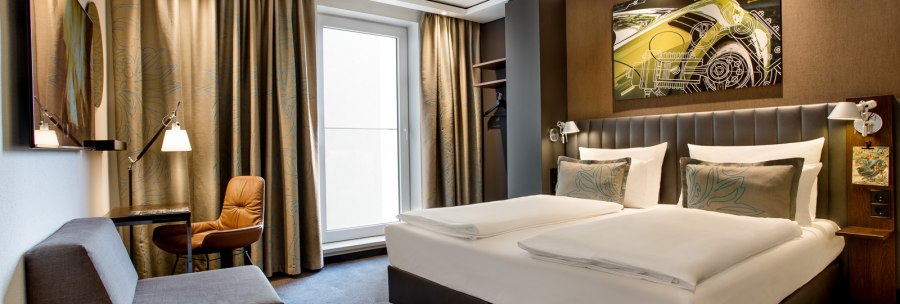 Modernes Hotelzimmer mit Doppelbett, Sofa, Schreibtisch und großem Fenster. An der Wand hängt ein Kunstwerk mit einem Auto-Motiv., © TOMAS Modernes Hotelzimmer mit Doppelbett, Sofa, Schreibtisch und großem Fenster. An der Wand hängt ein Kunstwerk mit einem Auto-Motiv., © TOMAS