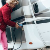 A woman in a red jacket and pink trousers connects a camper van to a power source. She is holding a blue cable in her hand., © Erlebnisregion Schwäbischer Albtrauf e. V. A woman in a red jacket and pink trousers connects a camper van to a power source. She is holding a blue cable in her hand., © Erlebnisregion Schwäbischer Albtrauf e. V.