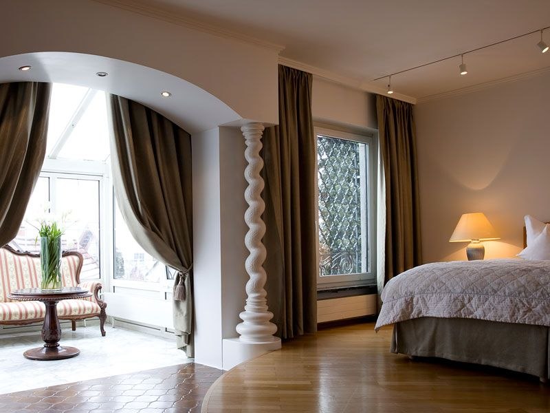 Elegantes Doppelzimmer mit einem großen Bett, Vorhängen, einem Sitzbereich mit Tisch und Stühlen sowie einer dekorativen Säule., © Romantisches Hotel Traube