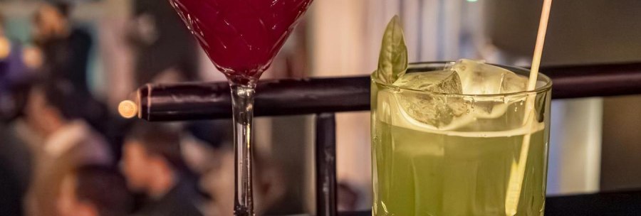 Zwei Cocktails auf einer Bar: ein roter im Martiniglas und ein gr&uuml;ner im Tumblerglas mit Strohhalm, im Hintergrund unscharfe Bar-Szene., &copy; SMG, Sarah Schmid