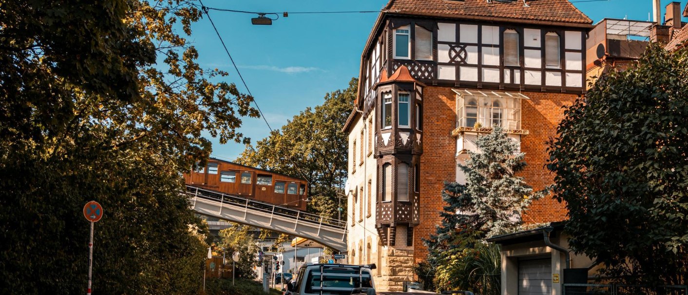 Eine historische Seilbahn fährt an einem Fachwerkhaus vorbei, umgeben von Bäumen und einem blauen Himmel., © Stuttgart Marketing GmbH, Sarah Schmid