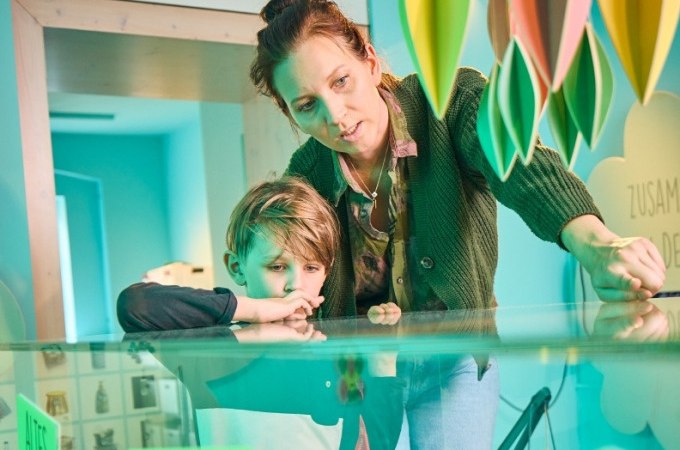 Eine Frau und ein Kind betrachten interessiert eine Ausstellung in einem Museum. Bunte Dekorationen hängen von der Decke., © Landesmuseum Württemberg