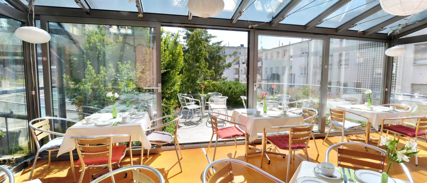 Ein heller Wintergarten mit gedeckten Tischen, roten und grünen Kissen, Blick auf Terrasse und umliegende Gebäude., © Abalon Hotel Ideal Ein heller Wintergarten mit gedeckten Tischen, roten und grünen Kissen, Blick auf Terrasse und umliegende Gebäude., © Abalon Hotel Ideal