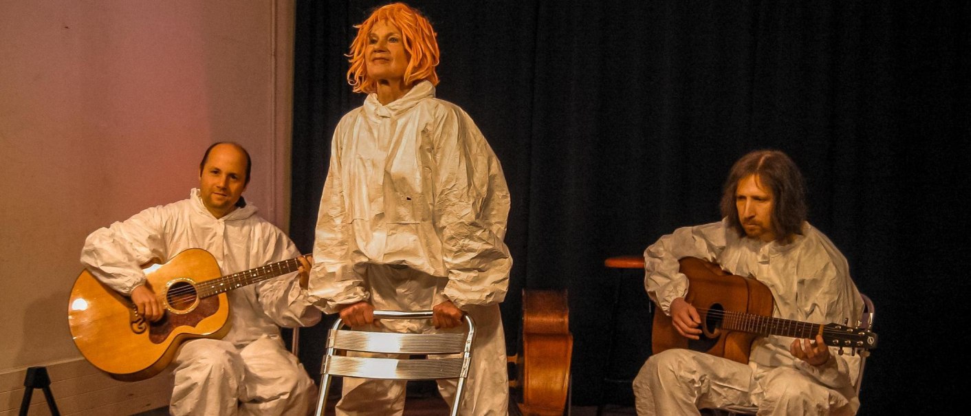 Drei Personen in weißen Anzügen auf einer Bühne. Zwei spielen Gitarre, eine steht mit orangefarbener Perücke und hält einen Stuhl., © Dreigroschentheater