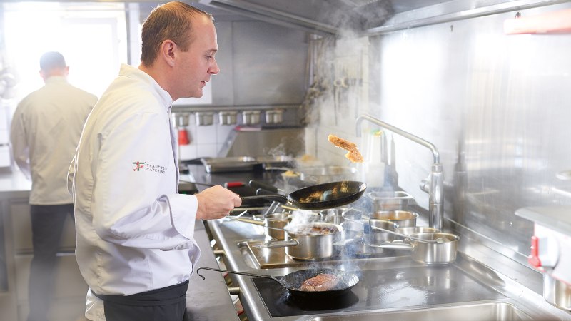 Ein Koch in weißer Uniform brät Fleisch in einer Pfanne in einer professionellen Küche. Im Hintergrund ist ein weiterer Koch zu sehen., © SMG, Jean-Claude Winkler