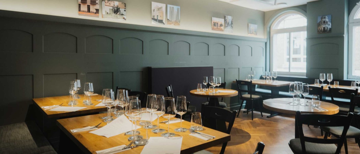 Ein stilvoll eingerichtetes Restaurant mit gedeckten Holztischen, Weingläsern und Bildern an der Wand. Große Fenster lassen Tageslicht herein., © Kati Guenter