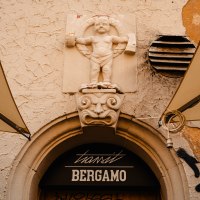 Stone relief of a child above an entrance with the inscription 'transit Bergamo'. The plaster is crumbling, graffiti and awnings are visible., &copy; Sarah Schmid