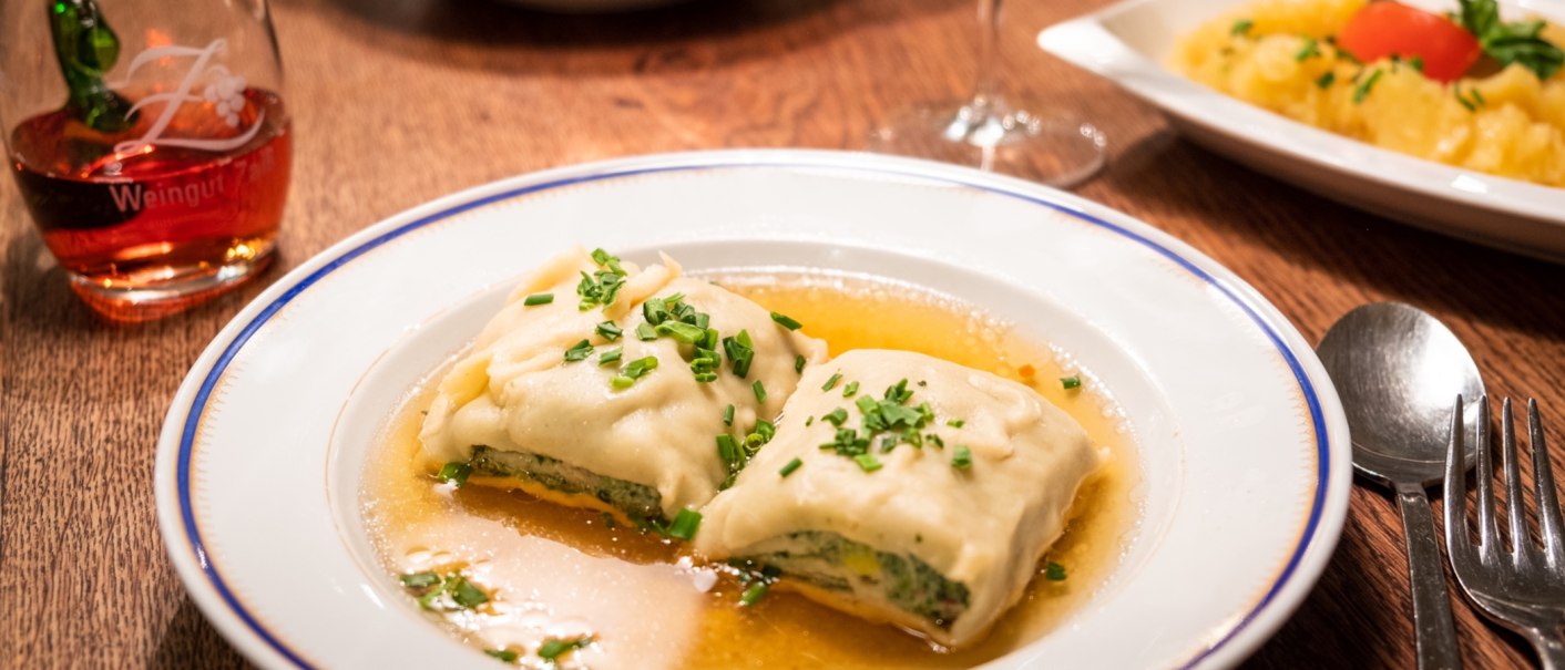 Two Maultaschen in broth on a plate, garnished with chives. Next to it, a glass of red wine and cutlery on a wooden table., © Weingut Zaiß