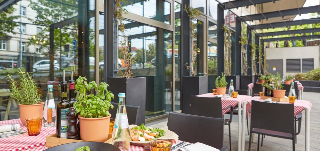 Restaurantterrasse mit rot-weiß karierten Tischdecken, Kräutertöpfen und gedeckten Tischen. Pasta und Getränke sind serviert., © Park Inn by Radisson Stuttgart