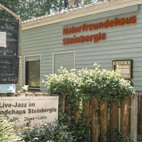 The NaturFreundeHaus Steinbergle with a sign announcing live jazz, surrounded by plants and a wooden fence., © NaturFreundehaus Steinbergle, Stuttgart