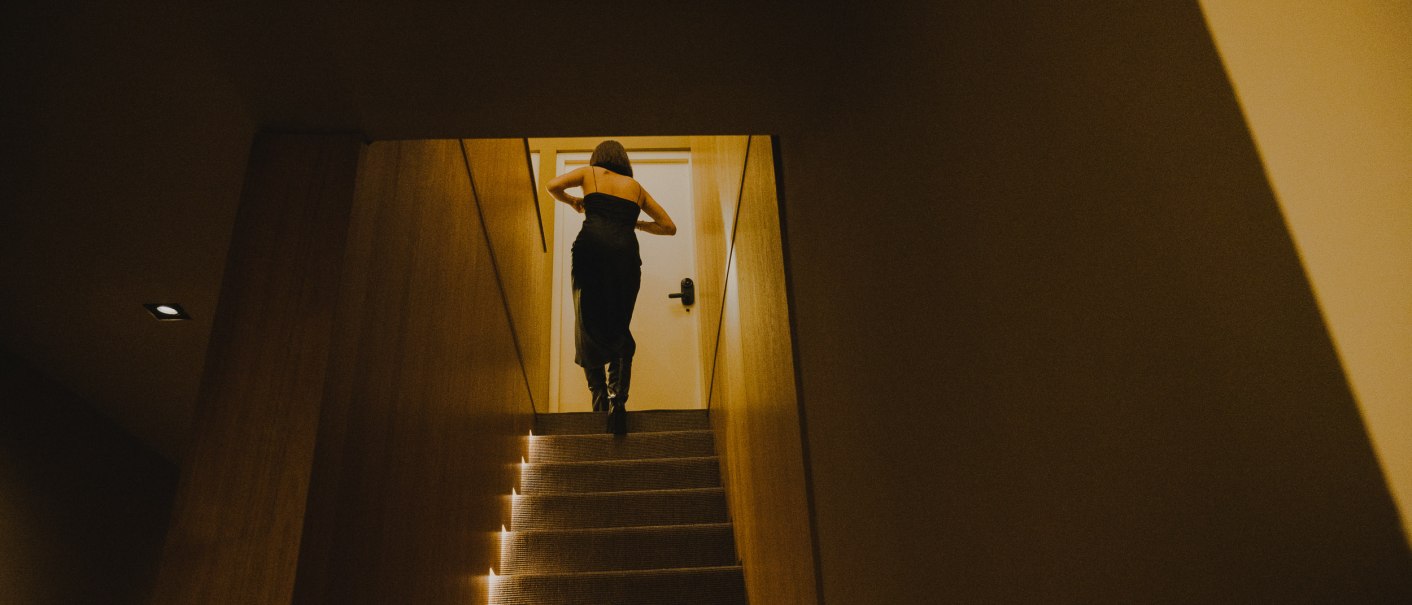 Eine Person in einem dunklen Kleid geht eine beleuchtete Treppe hinauf zu einer T&uuml;r in einem schmalen Flur., &copy; Peter Oliver Wolff / DAS EDITH