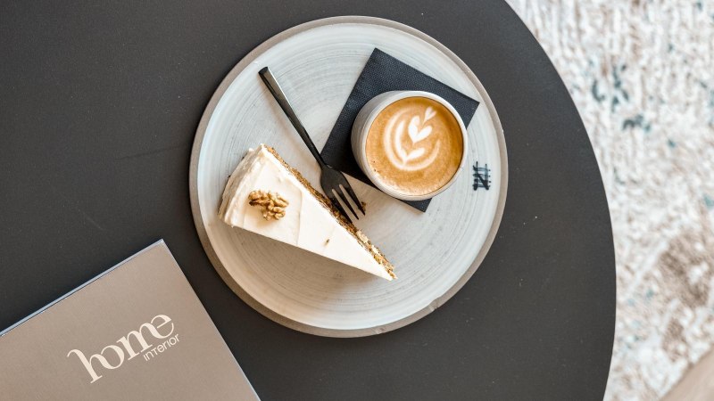 Cappuccino und Kuchen auf einem Teller auf einem schwarzen Tisch, daneben die Brosch&uuml;re &bdquo;home interior&ldquo;., &copy; Prosic GmbH - TARABA