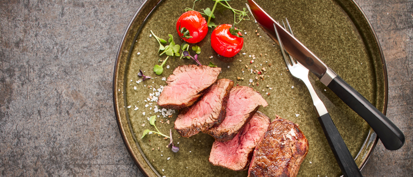 Ein Teller mit geschnittenem Steak, zwei Kirschtomaten und Besteck auf einem rustikalen Untergrund., &copy; MAREDO