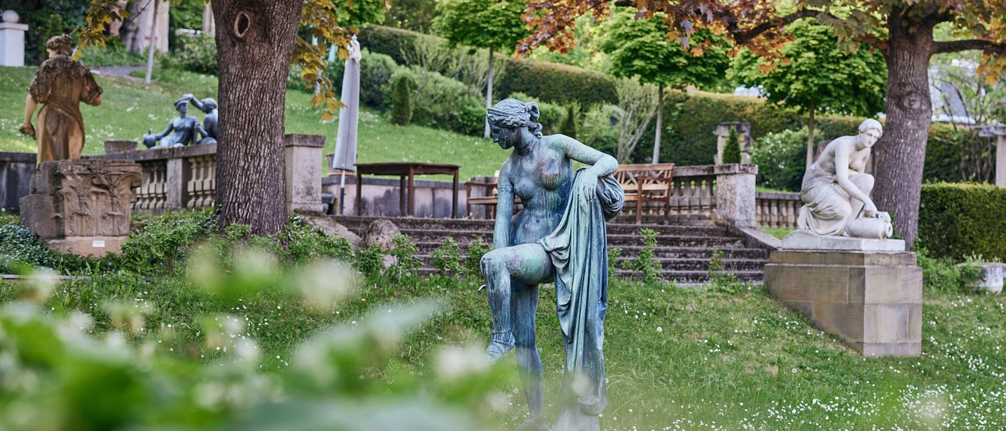 Ein Garten mit mehreren Skulpturen, umgeben von Bäumen und Grünflächen. Im Vordergrund eine bronzene Figur, im Hintergrund weitere Statuen., © Städtisches Lapidarium, Foto Julia Ochs
