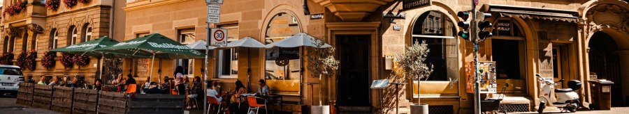 Street café with parasols in front of a historic building. People are sitting outside, enjoying the weather. Traffic signs and a Vespa are visible., © SMG Stuttgart Marketing GmbH - Sarah Schmid