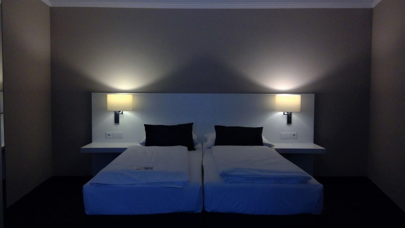 Zwei Einzelbetten mit weißen Laken und schwarzen Kissen in einem Hotelzimmer. An der Wand sind zwei Lampen angebracht, die warmes Licht spenden., © relexa Waldhotel Schatten Zwei Einzelbetten mit weißen Laken und schwarzen Kissen in einem Hotelzimmer. An der Wand sind zwei Lampen angebracht, die warmes Licht spenden., © relexa Waldhotel Schatten