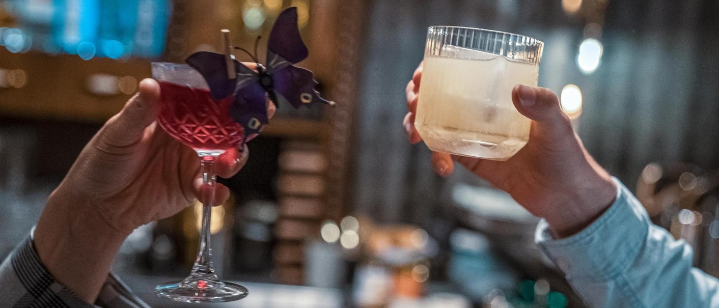 Zwei Hände stoßen mit Cocktails an, einer rot mit Schmetterlingsdeko, der andere klar mit Eiswürfel., © SMG, Martina Denker
