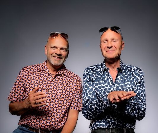 Zwei Männer in gemusterten Hemden und Jeans sitzen nebeneinander. Beide tragen Sonnenbrillen auf dem Kopf und lächeln vor einem grauen Hintergrund., © Theaterhaus Stuttgart e.V. Zwei Männer in gemusterten Hemden und Jeans sitzen nebeneinander. Beide tragen Sonnenbrillen auf dem Kopf und lächeln vor einem grauen Hintergrund., © Theaterhaus Stuttgart e.V.