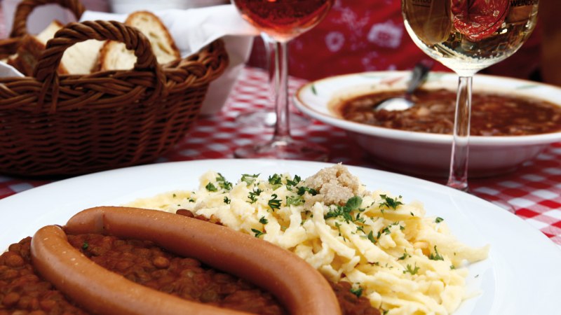 Ein Teller mit Linsen, Sp&auml;tzle und W&uuml;rstchen auf einem rot-wei&szlig; karierten Tischtuch. Im Hintergrund ein Brotkorb und ein Glas Wein., &copy; Stuttgart-Marketing GmbH